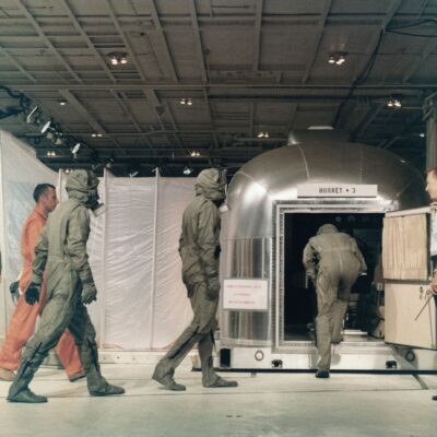 Astronaut Michael Collins enters the NASA MQF followed by Neil Armstrong and then "Buzz Aldrin." Behind Aldrin, in the orange flight suit, is NASA flight surgeon Bill Carpentier. To the left is the NASA Manager for the Apollo 11 quarantine program John Stonesifer, while holding the MQF door is Dr. Clarence Jernigan, who would become the NASA flight surgeon for Apollo 12. The sign over the door reads "Hornet + 3." This motto was selected by the Hornet CO, CAPT Carl Seiberlich, to establish a "safety first" mentality. The Hornet would return to Pearl Harbor with the full ship's complement, members of the media and other guests, plus 3 astronauts.

US Navy photo.