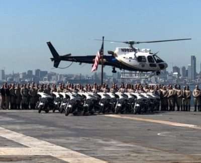 Helicopter hovering over CHP officers and motorcycles on deck with city skyline.