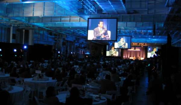 Large audience at formal event with multiple screens displaying speaker.