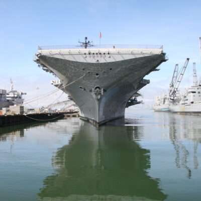 Aircraft carrier docked in a harbor, with cranes and ships in the background.