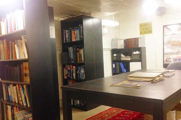 A room with a desk in the middle, filled with books and papers. There is a red carpet on the floor, and the walls are lined with bookshelves.