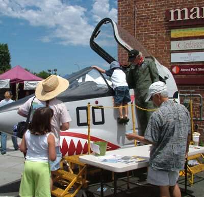 Children and adults exploring a fighter jet with shark mouth nose art at an outdoor event.