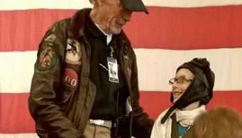 Elderly man in flight jacket talking with a child in pilot hat.