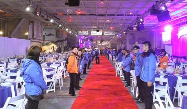 Event staff lining red carpet at formal dinner setup inside large venue.