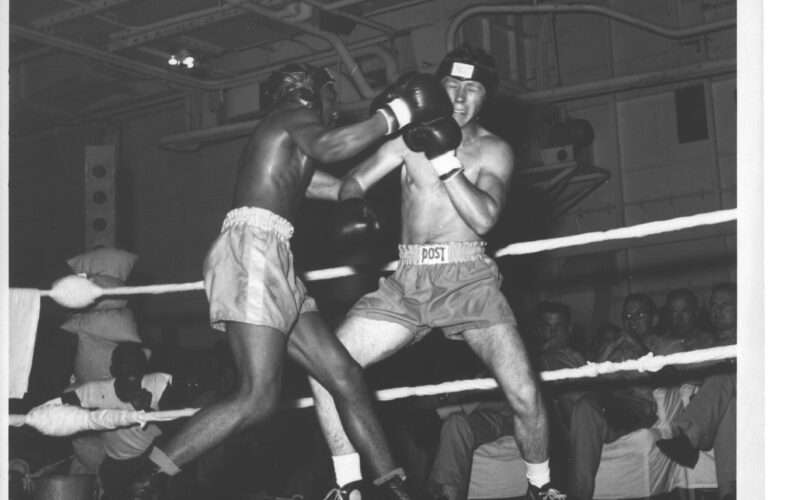 Boxing match between two men on USS Hornet, spectators watching, black and white photo.