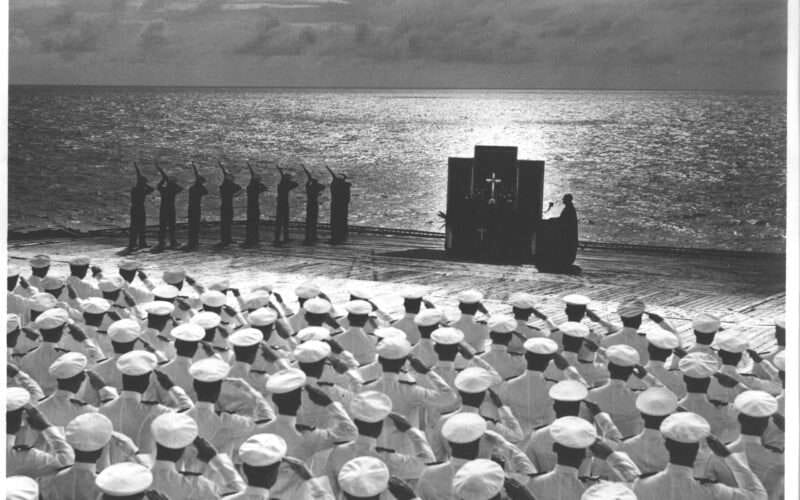 Navy personnel at ceremony on USS Hornet deck, sunset over the ocean, black and white photo.