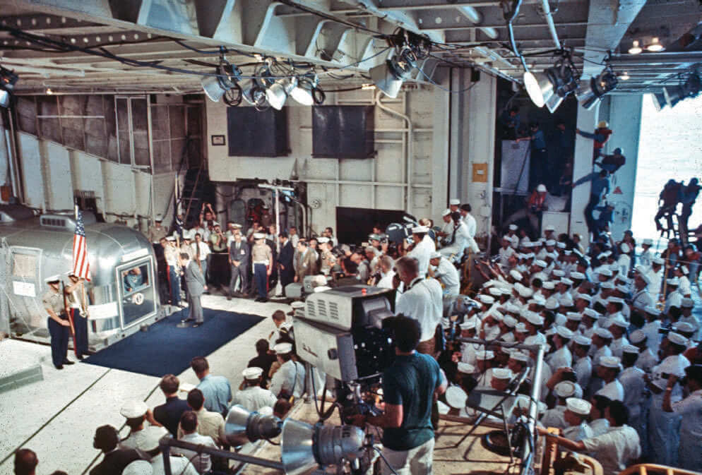 President Nixon with Apollo 11 astronauts in hangar deck, surrounded by Navy personnel and press.