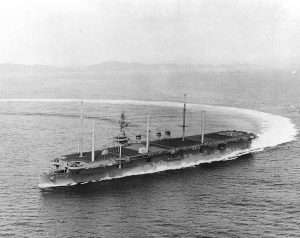 Aerial view of the USS Arlington (AGMR-2) making a sharp turn in the ocean with a coastline visible in the background.