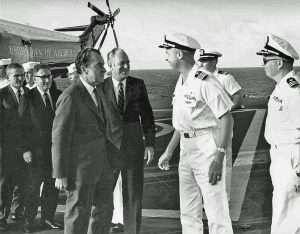 President Richard Nixon and other officials greeting naval officers on the deck of an aircraft carrier, with a helicopter marked 'United States of America' in the background.