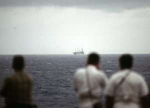 Three people in the foreground, blurred, looking out at a large ship in the distance on the ocean under a cloudy sky.